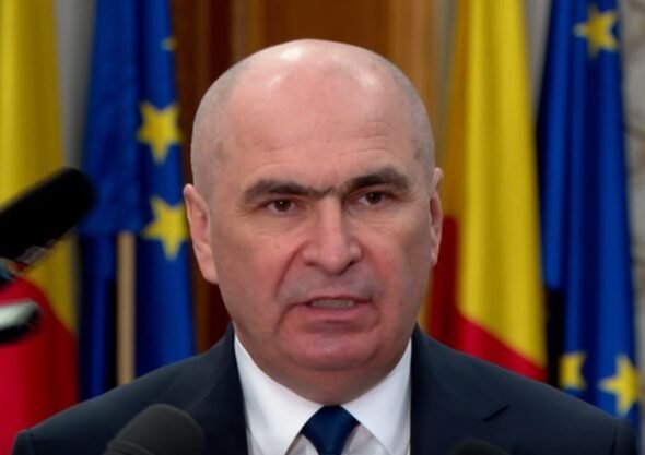 Bald man in a suit speaking at a press conference with microphones in front and flags in the background.