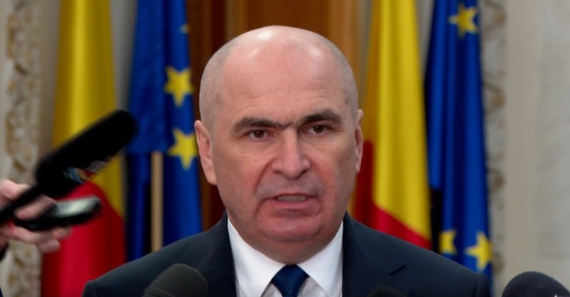 Bald man in a suit speaking at a press conference with microphones in front and flags in the background.