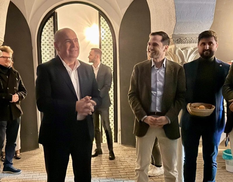 Group of people in a stone-arched hallway; a bald man in a dark suit speaks as a smiling man in a brown blazer stands nearby.