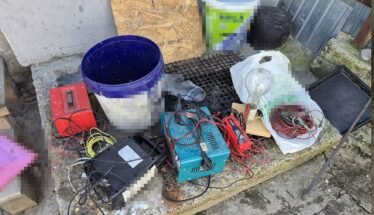 A cluttered assortment of electrical tools and wires on a stone ledge outside, including a blue bucket, battery charger, cables, and a plastic bag filled with small parts.
