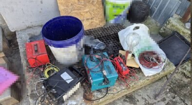 A cluttered assortment of electrical tools and wires on a stone ledge outside, including a blue bucket, battery charger, cables, and a plastic bag filled with small parts.