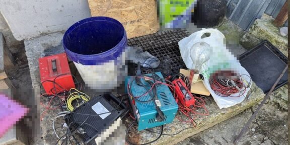 A cluttered assortment of electrical tools and wires on a stone ledge outside, including a blue bucket, battery charger, cables, and a plastic bag filled with small parts.