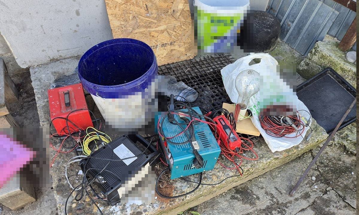 A cluttered assortment of electrical tools and wires on a stone ledge outside, including a blue bucket, battery charger, cables, and a plastic bag filled with small parts.