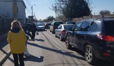 Pedestrians walk along a narrow street with parked cars on both sides; a woman in a bright yellow coat is in the foreground.