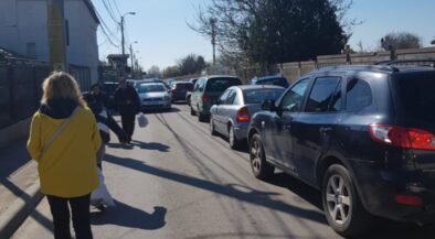 Pedestrians walk along a narrow street with parked cars on both sides; a woman in a bright yellow coat is in the foreground.