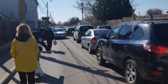 Pedestrians walk along a narrow street with parked cars on both sides; a woman in a bright yellow coat is in the foreground.