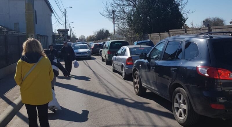 Pedestrians walk along a narrow street with parked cars on both sides; a woman in a bright yellow coat is in the foreground.