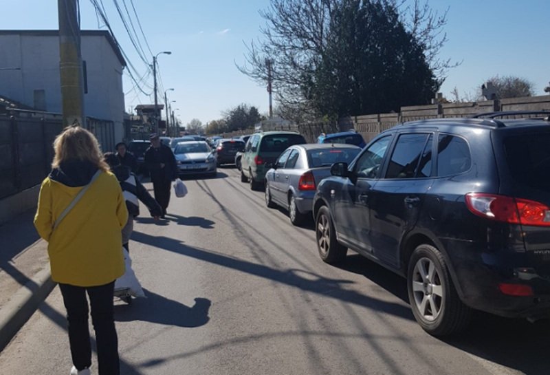 Pedestrians walk along a narrow street with parked cars on both sides; a woman in a bright yellow coat is in the foreground.