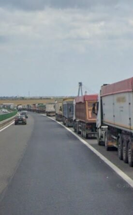 Row of large trucks lined up on the right shoulder of a highway, with a car in the left lane and open landscape beside the road.