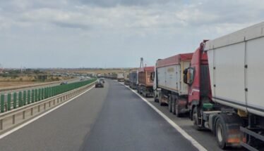 Row of large trucks lined up on the right shoulder of a highway, with a car in the left lane and open landscape beside the road.