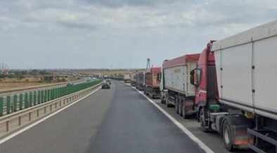 Row of large trucks lined up on the right shoulder of a highway, with a car in the left lane and open landscape beside the road.