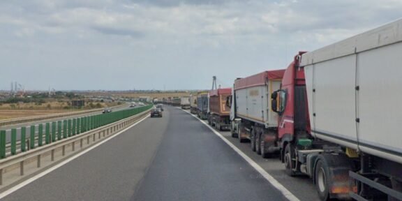 Row of large trucks lined up on the right shoulder of a highway, with a car in the left lane and open landscape beside the road.