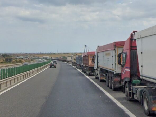 Row of large trucks lined up on the right shoulder of a highway, with a car in the left lane and open landscape beside the road.