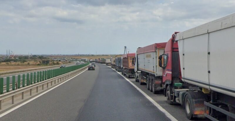 Row of large trucks lined up on the right shoulder of a highway, with a car in the left lane and open landscape beside the road.