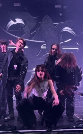 Five-person rock band posing on a stage with drums and guitars, under purple stage lights and abstract backdrop.