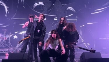 Five-person rock band posing on a stage with drums and guitars, under purple stage lights and abstract backdrop.