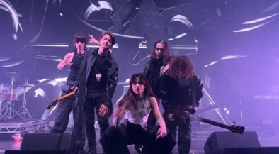 Five-person rock band posing on a stage with drums and guitars, under purple stage lights and abstract backdrop.