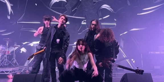 Five-person rock band posing on a stage with drums and guitars, under purple stage lights and abstract backdrop.