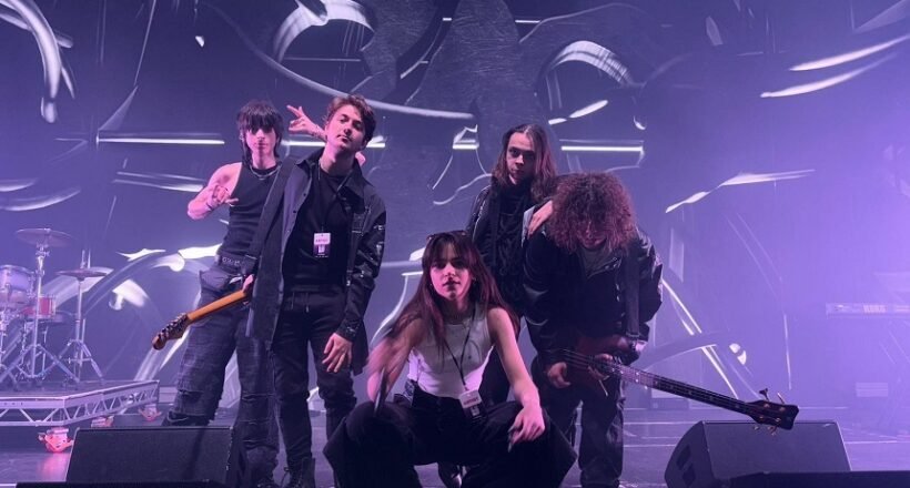 Five-person rock band posing on a stage with drums and guitars, under purple stage lights and abstract backdrop.