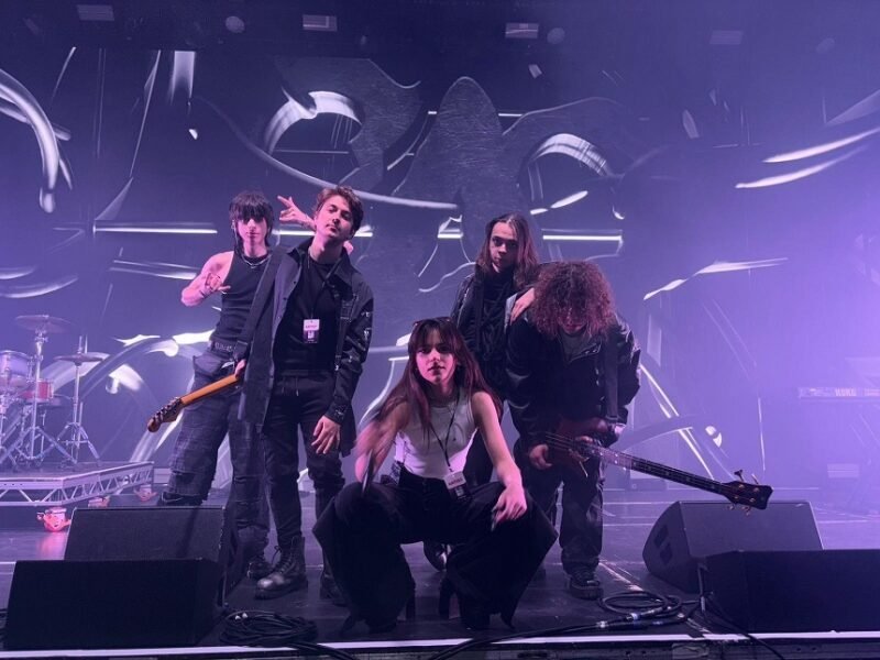 Five-person rock band posing on a stage with drums and guitars, under purple stage lights and abstract backdrop.