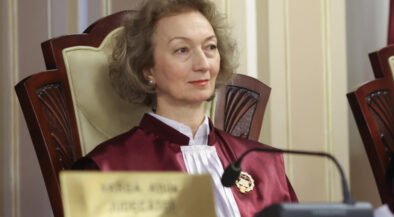 Female judge in a burgundy robe sits at a courtroom bench, looking slight to the right, with a nameplate and microphone in front of her.