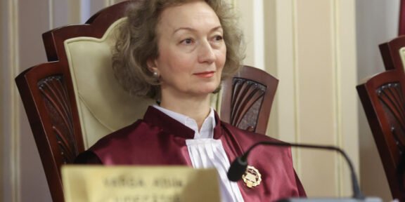 Female judge in a burgundy robe sits at a courtroom bench, looking slight to the right, with a nameplate and microphone in front of her.