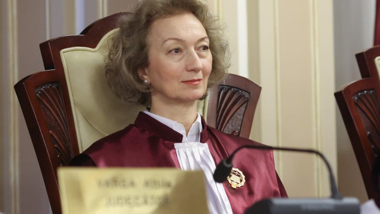 Female judge in a burgundy robe sits at a courtroom bench, looking slight to the right, with a nameplate and microphone in front of her.