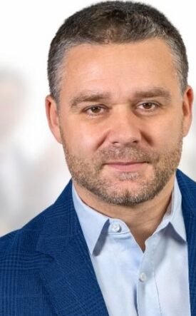 Professional portrait of a man in a blue blazer, foreground, with a crowd blurred in the background.