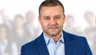 Professional portrait of a man in a blue blazer, foreground, with a crowd blurred in the background.