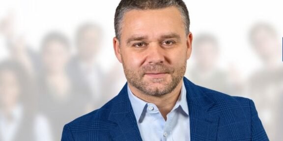 Professional portrait of a man in a blue blazer, foreground, with a crowd blurred in the background.