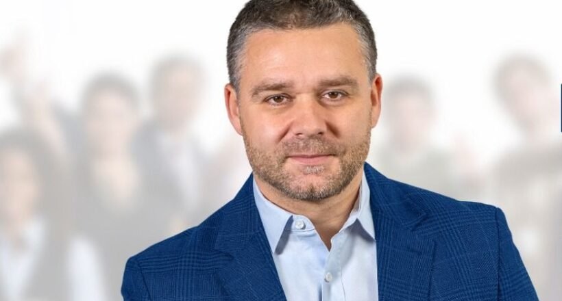 Professional portrait of a man in a blue blazer, foreground, with a crowd blurred in the background.