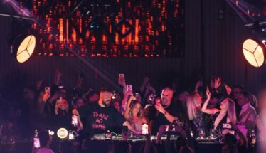 Crowd at a nightclub around a DJ booth, many people filming with phones under purple and red stage lights. Behind them a large LED screen emits orange patterns.