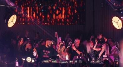 Crowd at a nightclub around a DJ booth, many people filming with phones under purple and red stage lights. Behind them a large LED screen emits orange patterns.