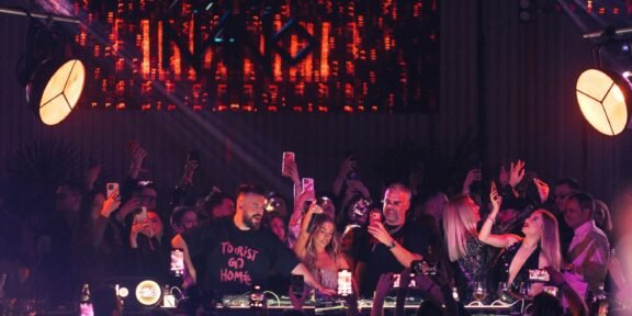 Crowd at a nightclub around a DJ booth, many people filming with phones under purple and red stage lights. Behind them a large LED screen emits orange patterns.