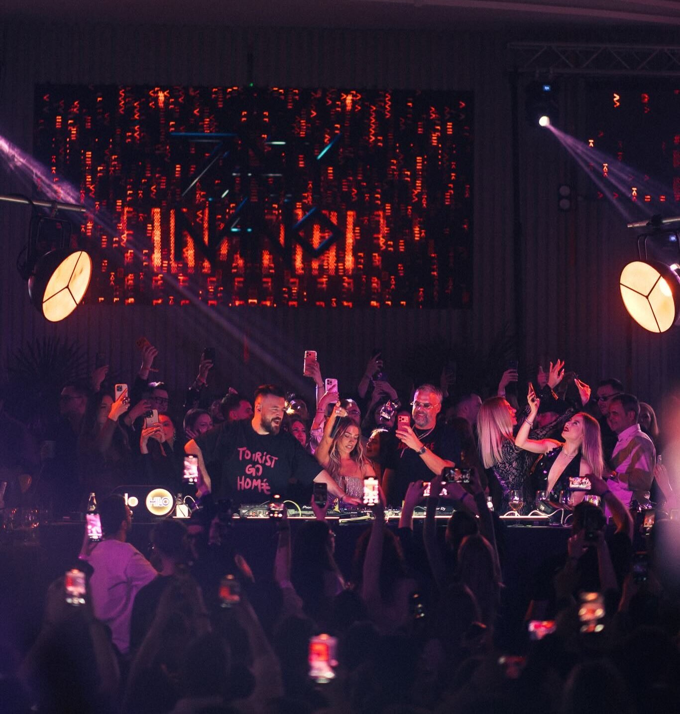 Crowd at a nightclub around a DJ booth, many people filming with phones under purple and red stage lights. Behind them a large LED screen emits orange patterns.