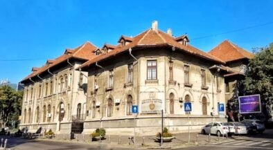 Historic beige educational building with arched windows and a red-tiled roof on a sunny city street, blue sky above.