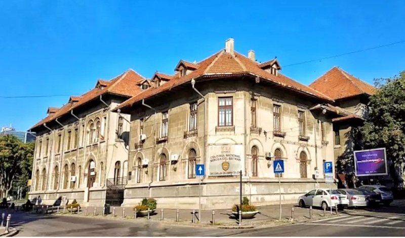 Historic beige educational building with arched windows and a red-tiled roof on a sunny city street, blue sky above.