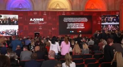 Indoor political event with a large red stage and multiple screens; a crowd seated facing the stage as speakers prepare to address them, banners read 'AJUNGE' and 'PSD' in the backdrop.