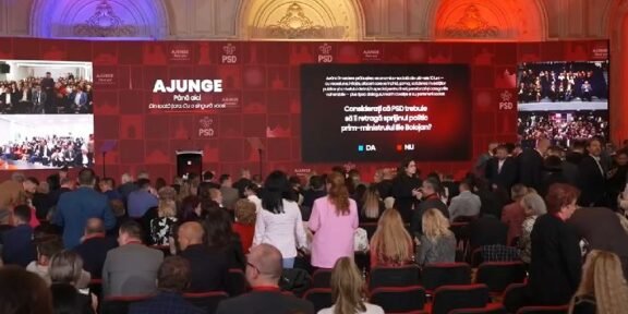 Indoor political event with a large red stage and multiple screens; a crowd seated facing the stage as speakers prepare to address them, banners read 'AJUNGE' and 'PSD' in the backdrop.