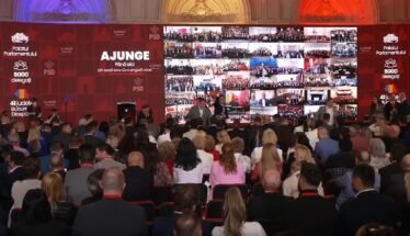 Large conference hall with a red backdrop; speakers on stage and a mosaic video wall displaying many small screens of attendees.