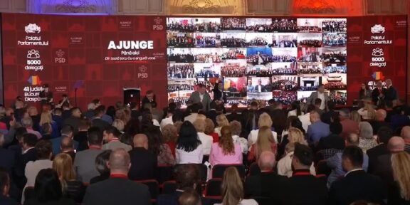 Large conference hall with a red backdrop; speakers on stage and a mosaic video wall displaying many small screens of attendees.