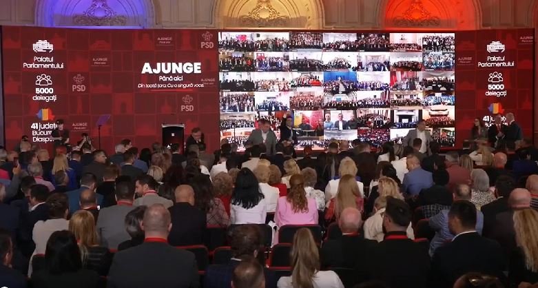 Large conference hall with a red backdrop; speakers on stage and a mosaic video wall displaying many small screens of attendees.