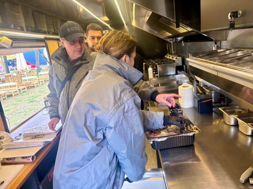 Inside a stainless steel food truck kitchen, a woman in a blue jacket places food from a foil pan while two men watch from behind.