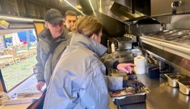 Inside a stainless steel food truck kitchen, a woman in a blue jacket places food from a foil pan while two men watch from behind.