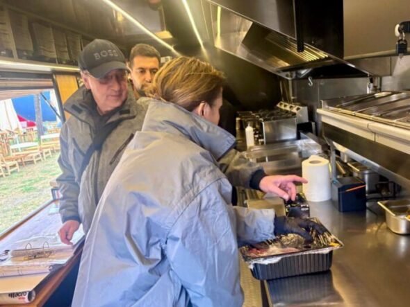 Inside a stainless steel food truck kitchen, a woman in a blue jacket places food from a foil pan while two men watch from behind.