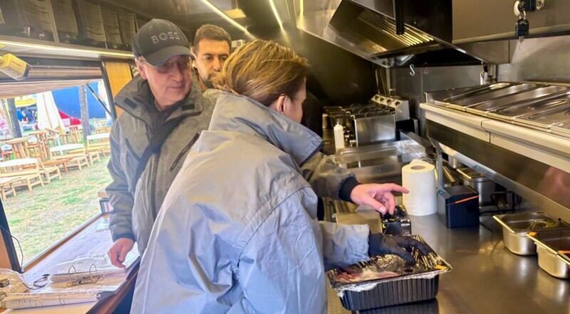 Inside a stainless steel food truck kitchen, a woman in a blue jacket places food from a foil pan while two men watch from behind.