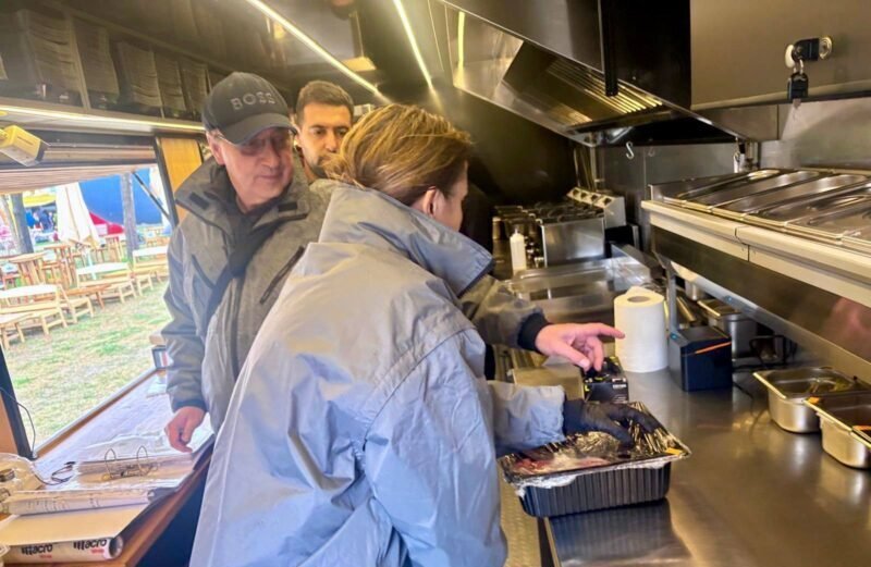 Inside a stainless steel food truck kitchen, a woman in a blue jacket places food from a foil pan while two men watch from behind.