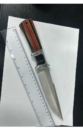 A metallic knife with a striped wooden handle resting on a sheet of white paper beside a ruler on a dark surface, viewed from above.
