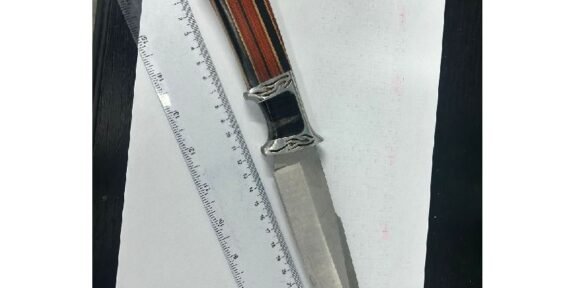 A metallic knife with a striped wooden handle resting on a sheet of white paper beside a ruler on a dark surface, viewed from above.