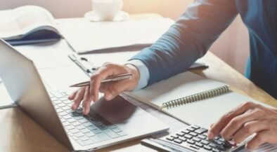 Person typing on a laptop while using a calculator and taking notes at a notebook on a sunny desk.